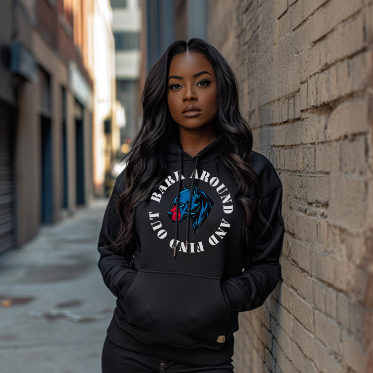 A woman stands in an urban alleyway, leaning against a brick wall and wearing a black hoodie with a bold graphic on the front. The design features a stylized blue and red dog's face, encircled by the text "Bark Around and Find Out." She gazes confidently at the camera, with long, wavy black hair and a serious expression. The background includes blurred building facades.